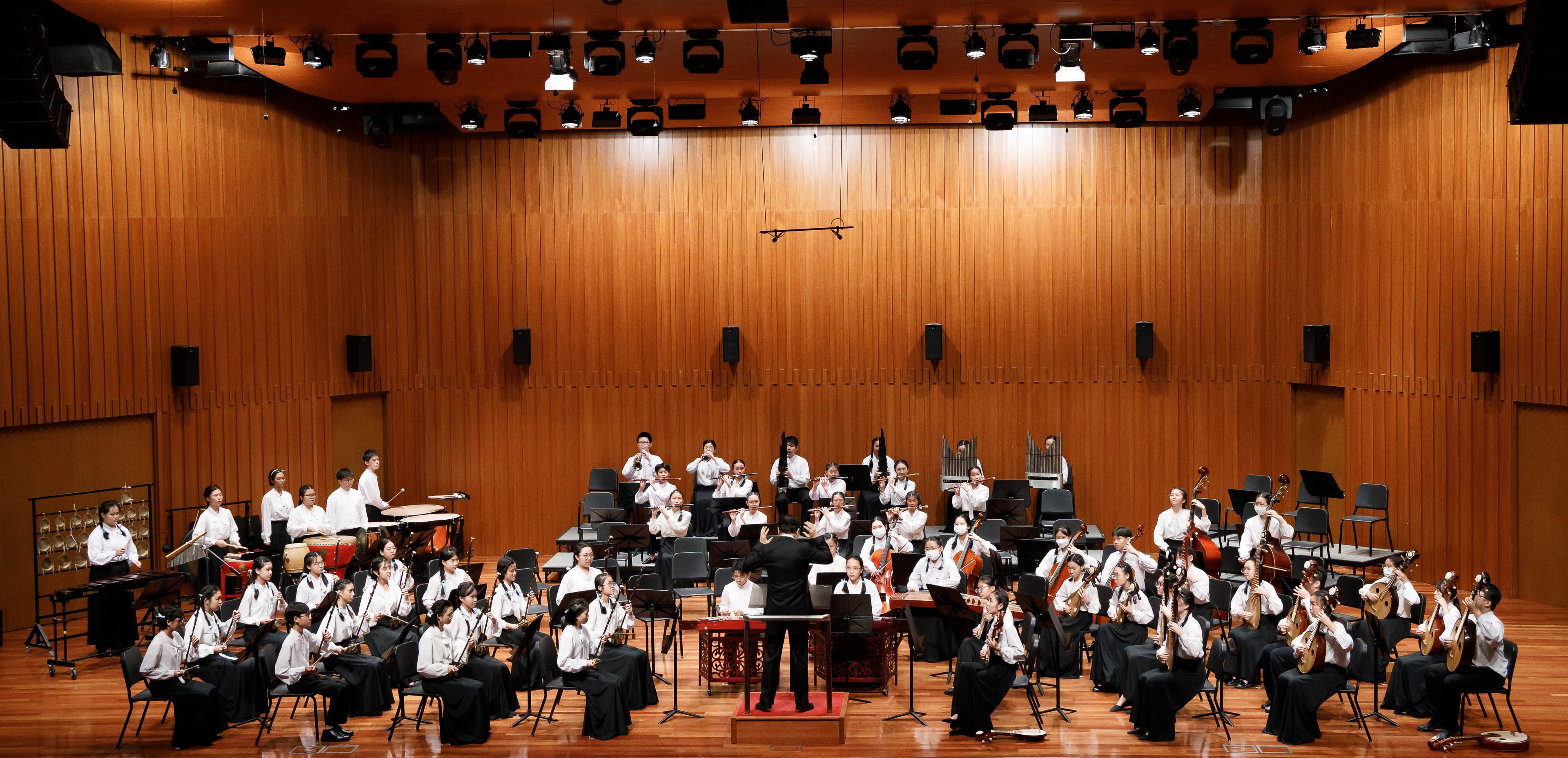 A conductor leads a large youth Chinese orchestra on a wooden-paneled concert hall stage.